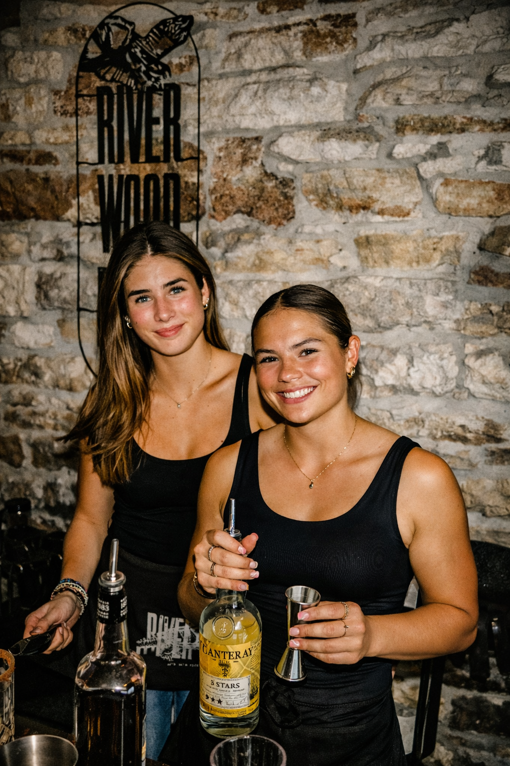 Two women holding a bottle and glass in front of a stone wall with 'River Wood' branding.