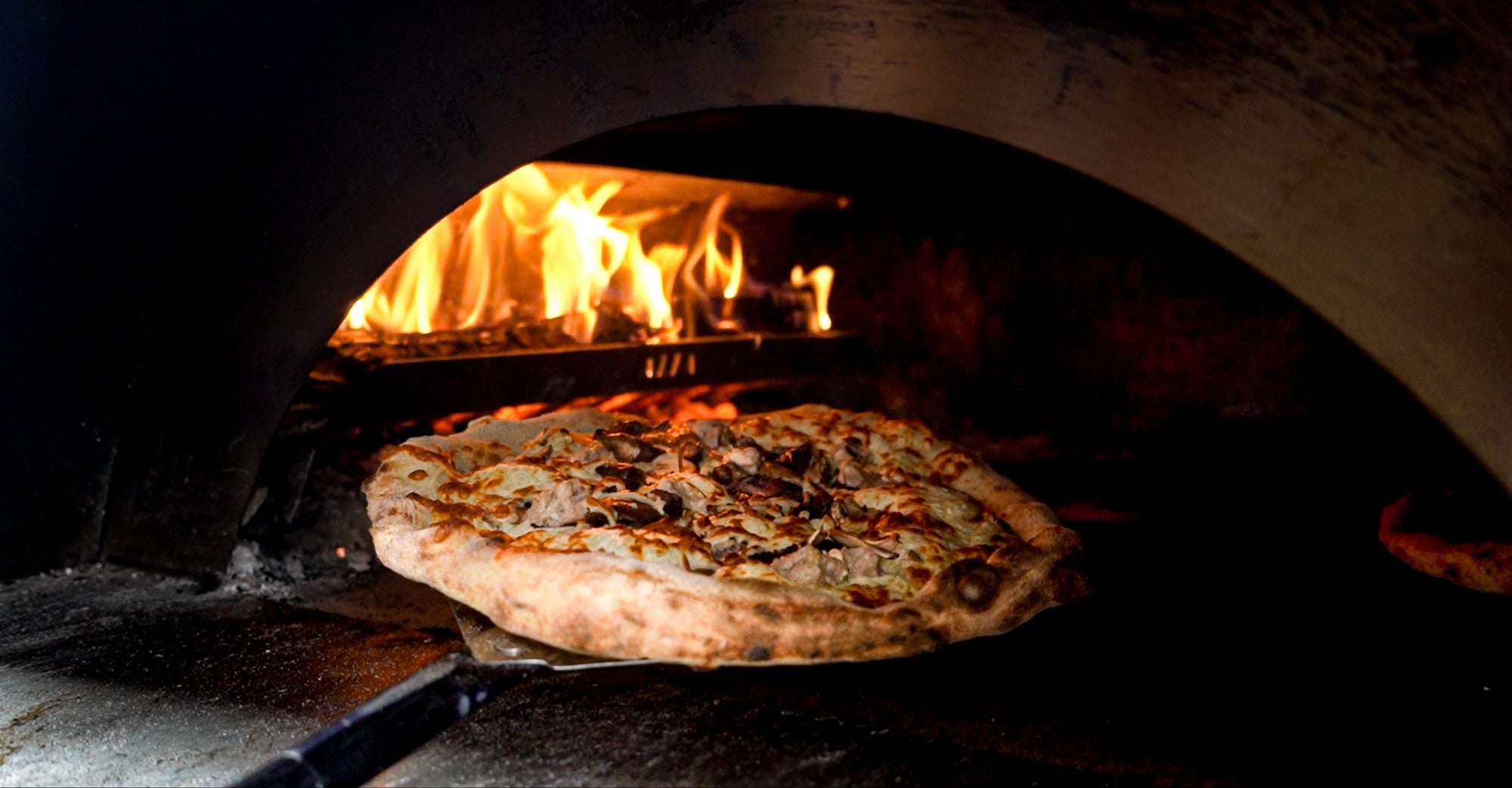 Pizza baking in a wood-fired oven with flames visible