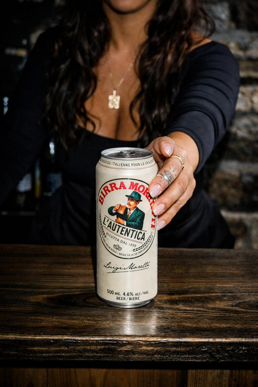 Person holding a Birra Moretti can on a wooden surface with a blurred background