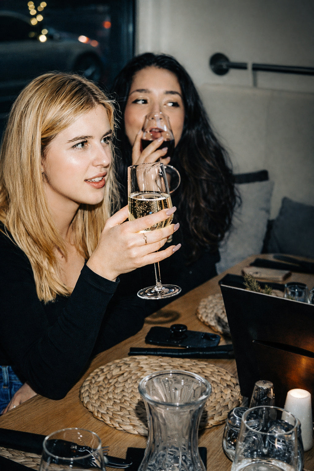 Two women at a table with drinks and a laptop, one holding a glass of champagne.